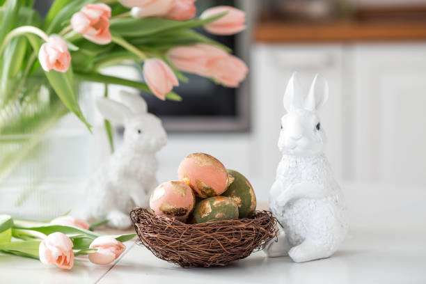 istock A bouquet of delicate tulips, Easter bunnies and eggs with a golden pattern on the table. There is a white kitchen in the background. Beautiful greeting card. The concept of minimalism. 2204203886