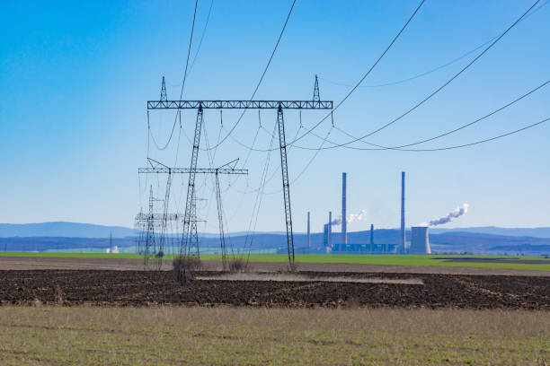 Wide shot of high voltage lines on a clear sunny through the green fields with a mountain in the distance. Coal power plant visible in the distance with smoke coming out of it.