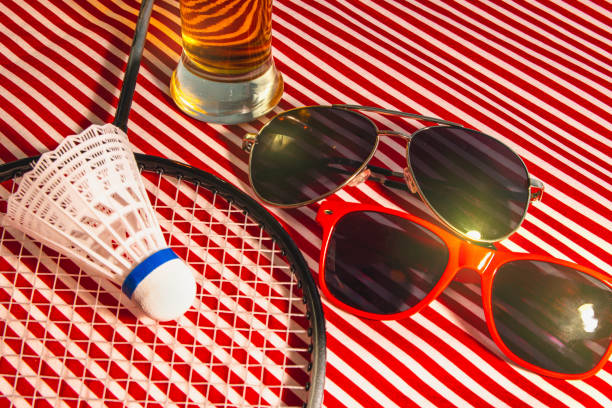 A pair of sunglasses and a racket are on a table. A glass of beer is also on the table. Scene is relaxed and casual, as it seems like someone is taking a break from playing badminton