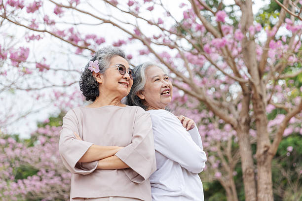 istock Two elderly women stand back-to-back, smiling joyfully under vibrant cherry blossom trees. 2204255574