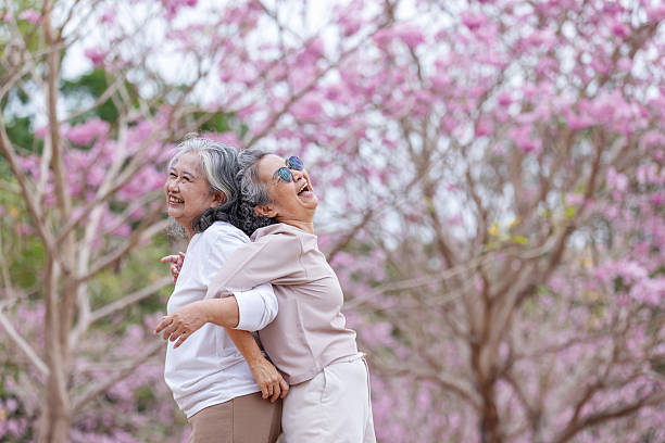 Two elderly women share a joyful moment, laughing and embracing under blooming cherry blossoms.