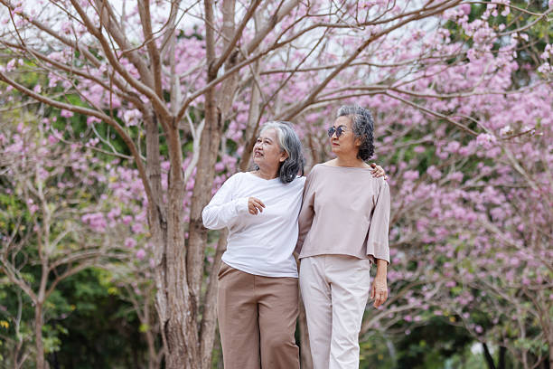istock Two elderly women walk arm in arm through a park filled with cherry blossoms, smiling joyfully. 2204255621