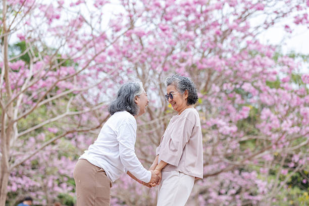 istock Two elderly women share a heartfelt moment under cherry blossoms, holding hands and laughing together. 2204255646