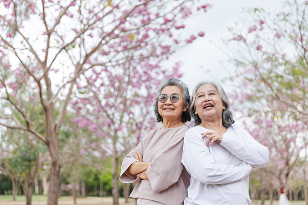 istock Two elderly women stand back-to-back, smiling joyfully under vibrant cherry blossom trees. 2204255704