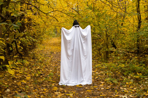 Trendy halloween celebration outdoors in autumn time. Handmade ghost costume from sheets, cap and glasses. Fall forest orange leaves.