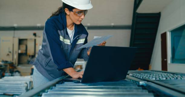 istock Young female engineer wearing hardhat uses laptop to analyze data and manage operations in factory. Robotic Industrial Factory. 2204259107