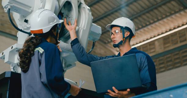 istock Two young engineers, one using laptop and the other handheld device, collaborate in factory to manage robotic arm. Robotic Industrial Factory. 2204259127