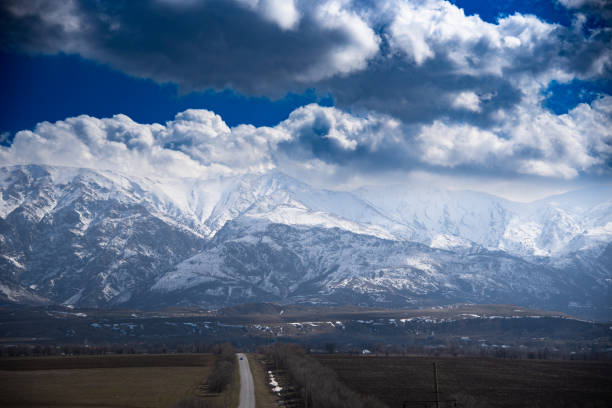 The image shows a majestic mountain range covered in snow under a dramatic sky filled with large, fluffy clouds. The mountains have a rugged and steep appearance, with various peaks and ridges visible. The sky above is a deep blue, contrasting beautifully with the white snow and the dark, shadowed areas of the mountains. The scene is serene and awe-inspiring, capturing the raw beauty of nature.