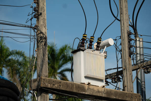 An electric transformer sits atop a utility pole, showcasing various wires and cables against a bright blue sky in a residential area.