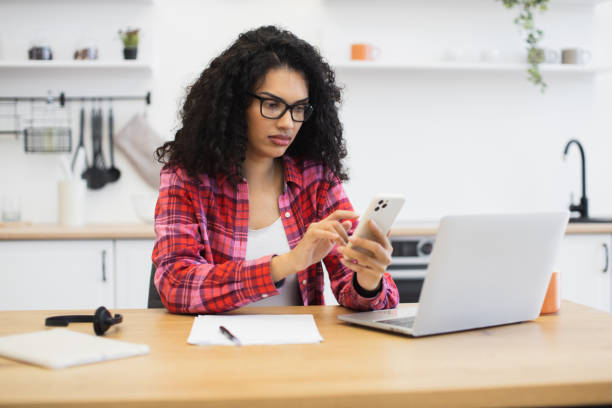 istock Focused young woman using smartphone while working on laptop in kitchen 2204271314