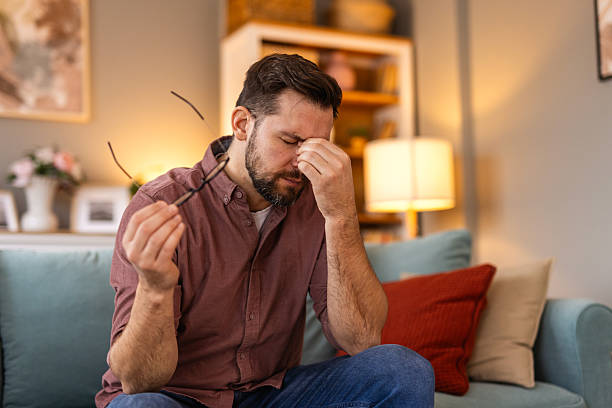 A stressed man sitting on a couch, holding his glasses and rubbing his forehead, overwhelmed with exhaustion or frustration.