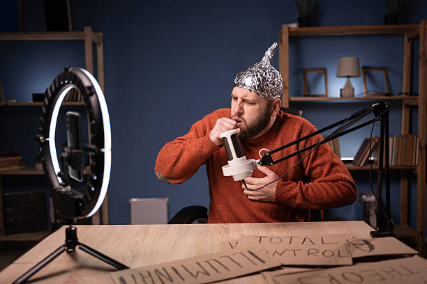 istock Paranoid man wearing foil hat recording video in home studio sickness suffering cough. Sitting at desk with ring lamp and smartphone, sharing theories, creating content for social media 2204276175