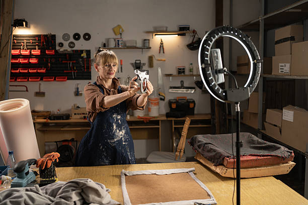 istock Middle aged craftswoman filming a tutorial about restoring furniture in her workshop using smartphone and ring lamp. She showing stapler for furniture upholstery. 2204276716
