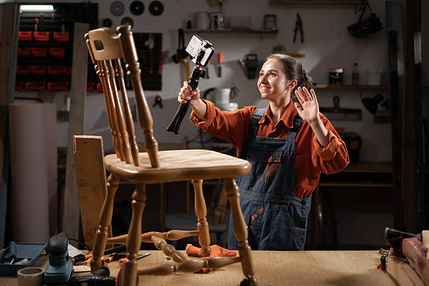 istock Female carpenter holding camera on tripod while recording diy video blog, she restoring old furniture in her workshop 2204276825