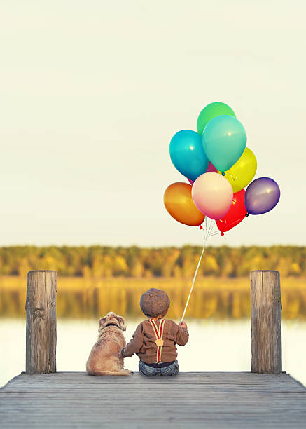 cute little boy with dog and colorfull balloons is sitiing on jetty