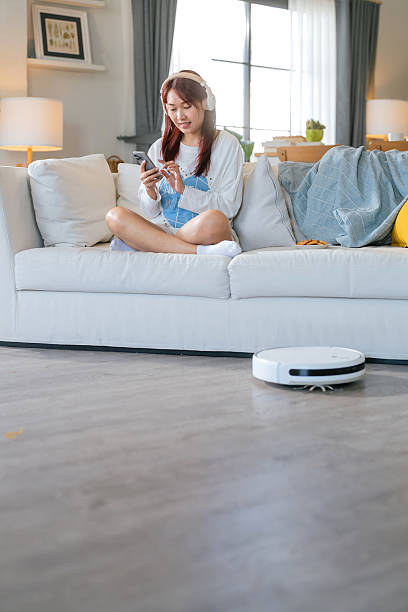 istock Young Asian woman relaxing sit on sofa home and turning on her smart vacuum cleaner using smartphone control robot while listening music headphone smart life smart technology lifestyle home hygiene 2204280376