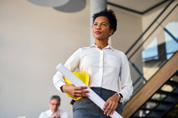 istock Confident female architect holding blueprints and hardhat in office 2204288236