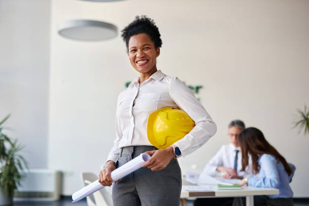 istock Confident female architect holding blueprints and yellow helmet in office 2204288241