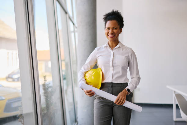 istock Confident female architect holding blueprints and hardhat in office 2204288242
