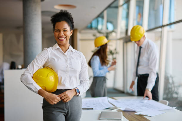istock Confident female architect holding yellow helmet in office with blueprint on table 2204288484