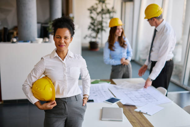 istock Confident female architect holding hardhat with colleagues working in background 2204288494