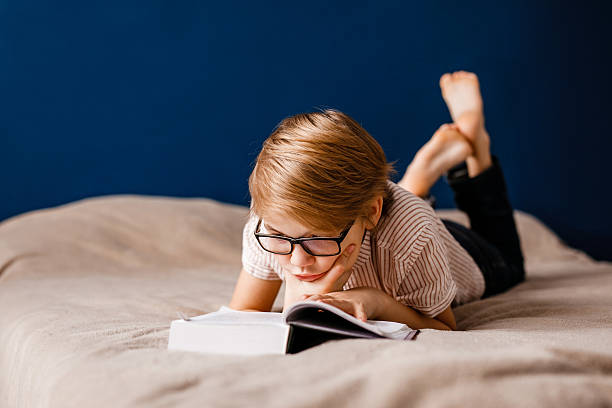 istock A 10-year-old boy with glasses is lying on the bed reading a big book 2204289583