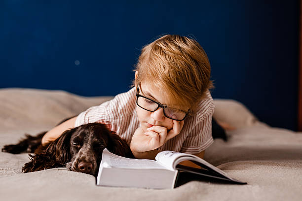 istock Boy with glasses lying in bed reading with his dog by his side 2204289679