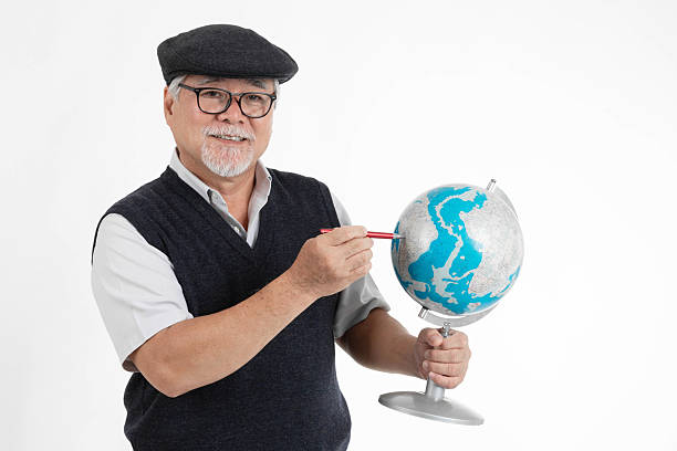 Senior man with glasses and flat cap smiling while pointing at a globe with a pencil, isolated on white background, concept of geography, travel planning, education, world exploration, and teaching