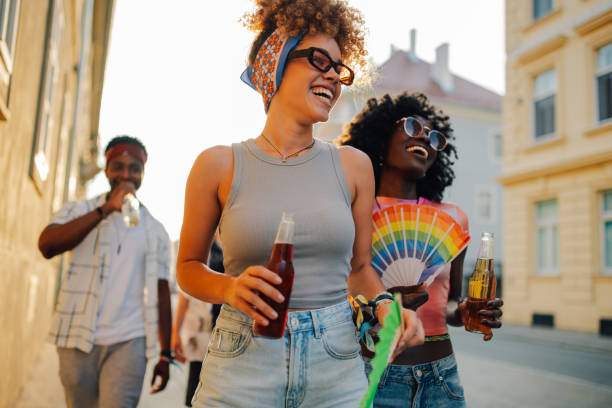 Two cheerful women holding colorful rainbow hand fans and drinks, walking and laughing joyfully with friends during a vibrant gay pride parade on a lively city street