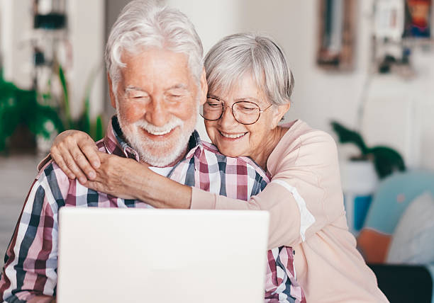 istock Mature white haired senior husband and wife sitting at home sharing internet connection using laptop, smiling and lovely modern up-to-date senior couple enjoying tender family moments together 2204311600