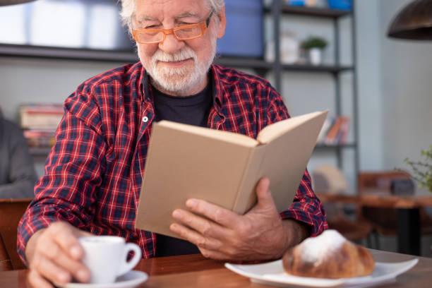 istock Smiling relaxed senior bearded man sitting at cafe table reading a book enjoying breakfast with coffee and croissant. Elderly caucasian male in eyeglasses and checkered shirt 2204318714