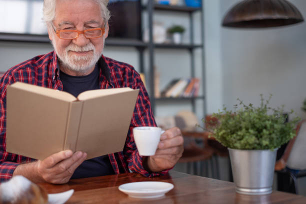 istock Smiling relaxed senior bearded man sitting at cafe table reading a book enjoying breakfast with coffee and croissant. Elderly caucasian male in eyeglasses and checkered shirt 2204318890