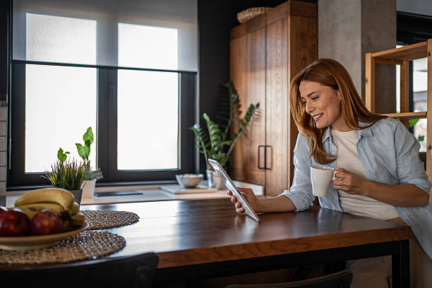 istock Smiling woman using digital tablet and drinking coffee in kitchen 2204342357