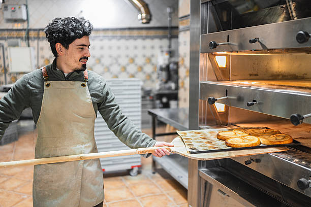 Baker using a wooden peel for placing a tray of freshly made palmiers into a professional oven, showcasing culinary skill and craftsmanship