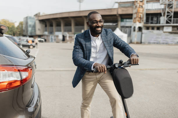 A smiling businessman in a formal outfit rides an electric scooter in an urban area, showcasing the modern blend of business and mobility in city life.