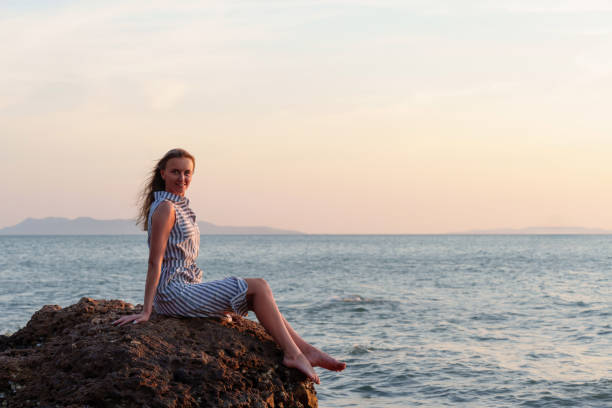 Young woman is sitting on a rock by the sea, enjoying the sunset, feeling the peace and quiet of nature, embracing the solitude and the beauty of the moment, during her solo travel adventure
