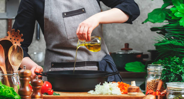 istock Chef pours olive oil in cast iron saucepan. Cozy kitchen with wooden table, kitchenware, vegetables, herbs and ingredients for cooking. Healthy food, eating, culinary, recipes, food blogging 2204402044