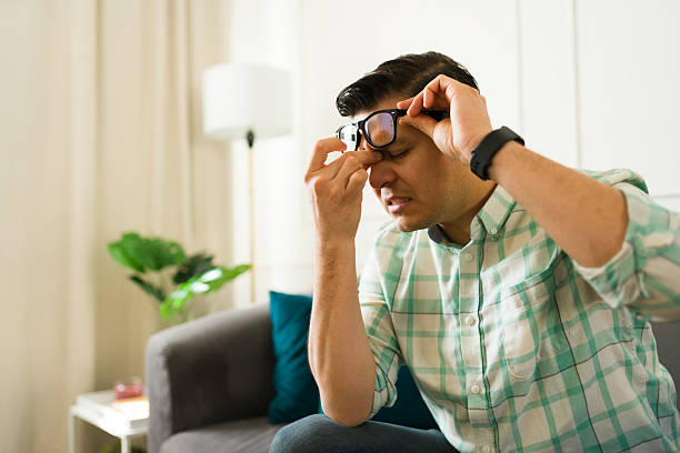 Frustrated hispanic male removing glasses, pressing nose bridge while experiencing intense headache on living room couch