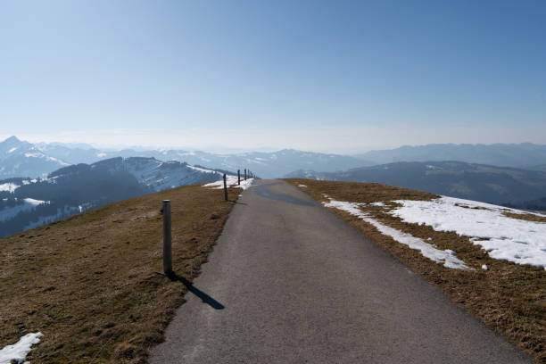 Paved hiking trail along the Swiss Hochalp mountain ridge with wooden guideposts, surrounded by melting snow and dormant grass. The path offers breathtaking views of distant snow-capped mountain ranges under a clear blue sky in early spring conditions.