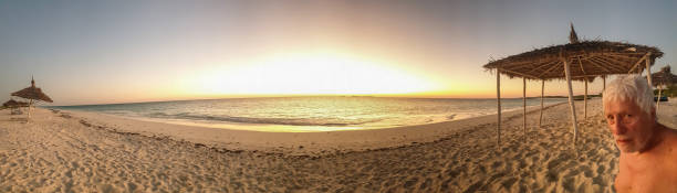 Panoramic view on sandy beach, straw umbrellas and deck chairs. At side senior men, head and shoulders, enjoying the time, Madagascar