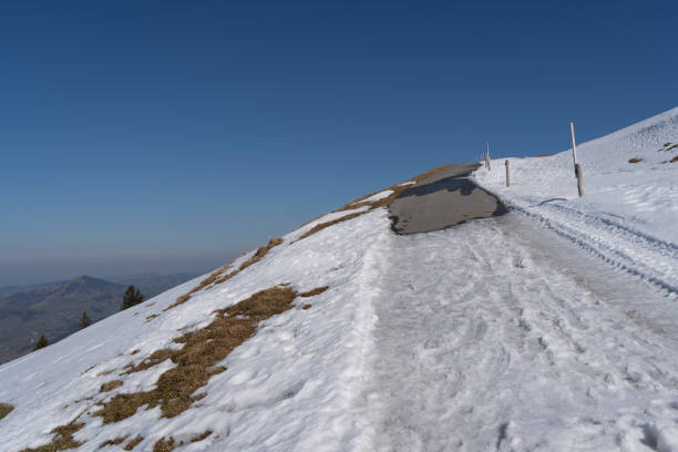 A paved mountain road curves between snow-covered slopes with wooden marker posts. The snow is melting to reveal patches of brown grass. Below, distant Alpine mountains fade into the horizon under a deep blue, cloudless sky. Tire tracks in the snow indicate recent passage along this high-altitude route.