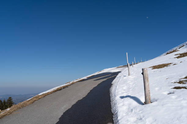 A paved mountain road curves between snow-covered slopes with wooden marker posts. The snow is melting to reveal patches of brown grass. Below, distant Alpine mountains fade into the horizon under a deep blue, cloudless sky. Tire tracks in the snow indicate recent passage along this high-altitude route.