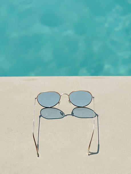 A pair of sunglasses rests casually on the poolside, reflecting the bright sun. The water shimmers beside them, adding to the relaxed, summery vibe of the scene.