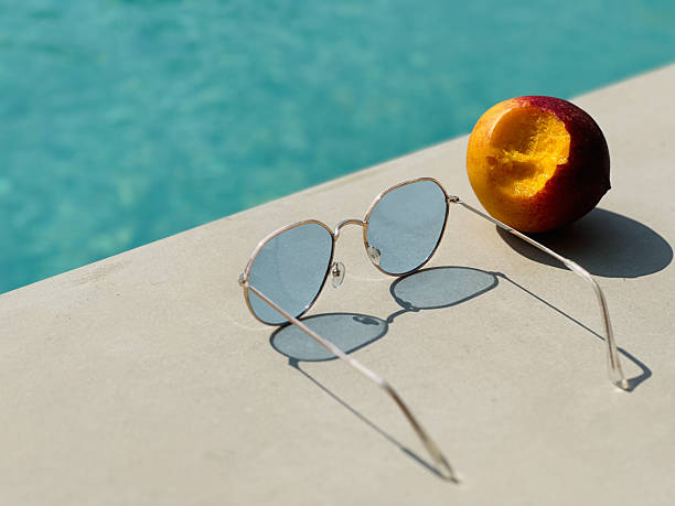 A ripe peach with a bite missing rests on the pool edge next to sunglasses. The sunlight creates a delicate shadow from the sunglasses on the pool’s surface, adding to the warm, inviting summer scene.
