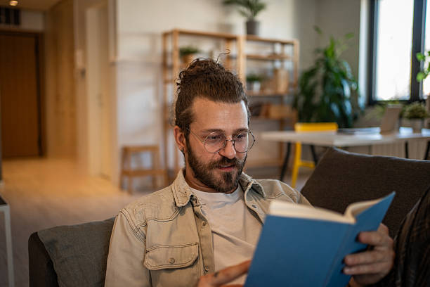 istock Young man reading a book on the sofa at home 2204427398