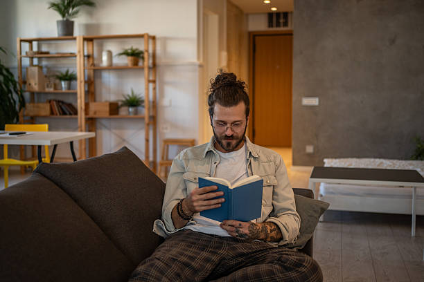 istock Young man reading a book on the sofa at home 2204427414