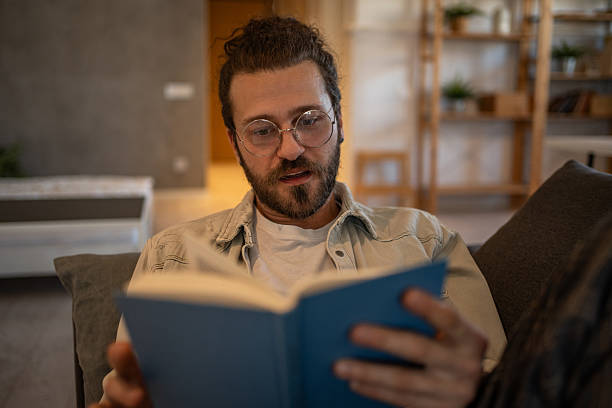 istock Young man reading a book on the sofa at home 2204427415