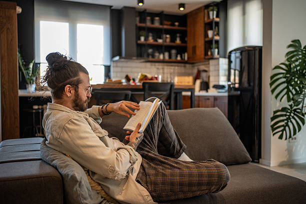 istock Young man reading a book on the sofa in his modern apartment 2204427432