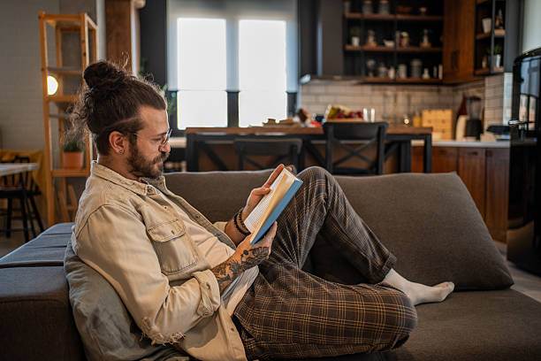 istock Young man reading a book on the sofa in his modern apartment 2204427440