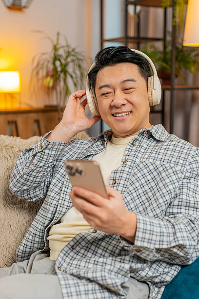 istock Happy overjoyed young man in wireless headphones using smartphone relaxing sitting on sofa at home 2204435598